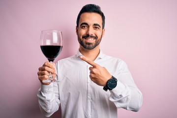 Young handsome man with beard drinking glass of red wine over isolated pink background very happy pointing with hand and finger