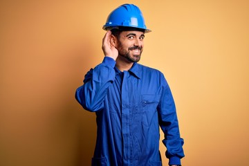 Mechanic man with beard wearing blue uniform and safety helmet over yellow background smiling with...
