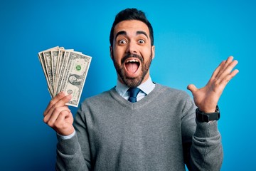 Young handsome man with beard holding bunch of dollars banknotes over blue background very happy and excited, winner expression celebrating victory screaming with big smile and raised hands