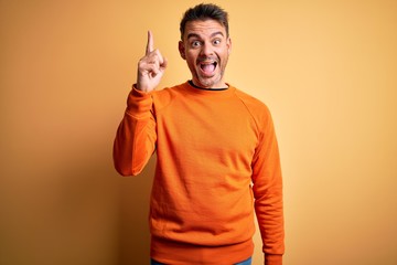 Young handsome man wearing orange casual sweater standing over isolated yellow background pointing finger up with successful idea. Exited and happy. Number one.