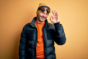 Young handsome skier man skiing wearing snow sportswear using ski goggles smiling positive doing ok sign with hand and fingers. Successful expression.