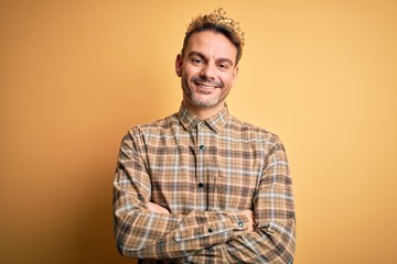 Young handsome man wearing golden crown of king over isolated yellow background happy face smiling with crossed arms looking at the camera. Positive person.