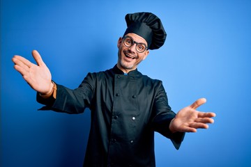Young handsome chef man wearing cooker uniform and hat over isolated blue background looking at the camera smiling with open arms for hug. Cheerful expression embracing happiness.
