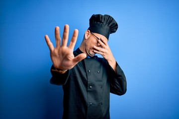 Young handsome chef man wearing cooker uniform and hat over isolated blue background covering eyes with hands and doing stop gesture with sad and fear expression. Embarrassed and negative concept.