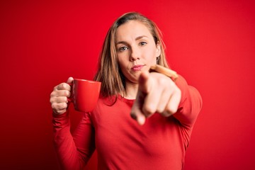 Young beautiful blonde woman drinking cup of coffee standing over isolated red background pointing with finger to the camera and to you, hand sign, positive and confident gesture from the front