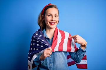 Young beautiful blonde woman wearing United States flag over isolated blue background with surprise face pointing finger to himself