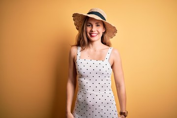 Young beautiful blonde woman on vacation wearing summer hat over yellow background with a happy and cool smile on face. Lucky person.