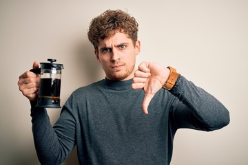 Young blond man with curly hair making coffee using coffemaker over white background with angry...