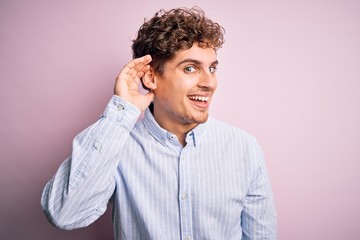 Young blond handsome man with curly hair wearing striped shirt over white background smiling with hand over ear listening an hearing to rumor or gossip. Deafness concept.