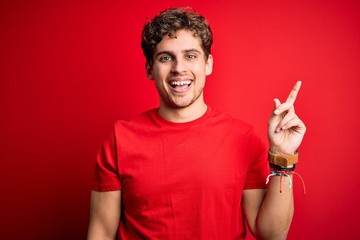 Fototapeta premium Young blond handsome man with curly hair wearing casual t-shirt over red background with a big smile on face, pointing with hand finger to the side looking at the camera.