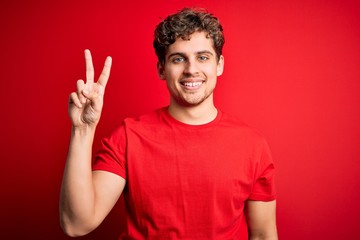 Naklejka premium Young blond handsome man with curly hair wearing casual t-shirt over red background smiling looking to the camera showing fingers doing victory sign. Number two.