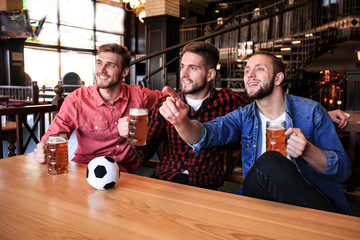 Fototapeta na wymiar Three men in casual clothes are cheering for football and holding bottles of beer while sitting at bar counter in pub.