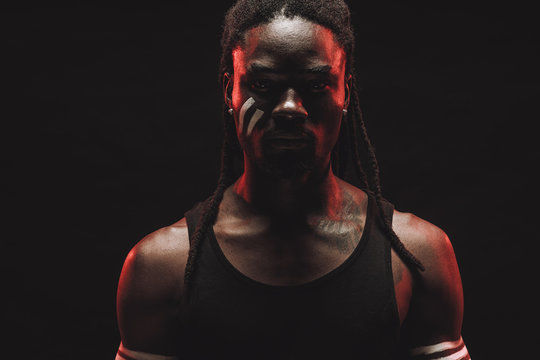 Confident, Serious Black Man With Dreadlocks Isolated Over Black Background. Young Muscular Man Stand In Black Shirt, With White Strips On Face, Confidently Look At Camera. Ethnic Models Concept