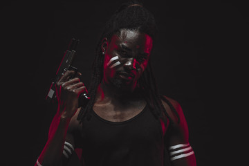 african ruffian man with steel arms isolated over black, furious hoodlum conceived to commit something criminal, looking at camera thoughtfully holding loaded gun, with red neon rays on body and face