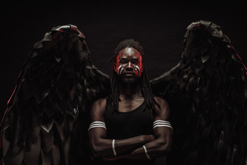 dark african angel with big black wings isolated, young serious muscular man wearing big wings on...