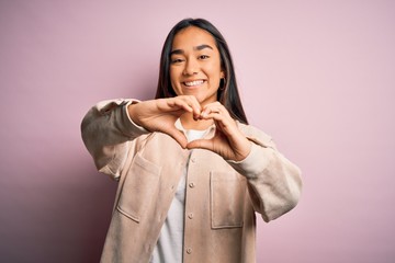 Young beautiful asian woman wearing casual shirt standing over pink background smiling in love doing heart symbol shape with hands. Romantic concept.