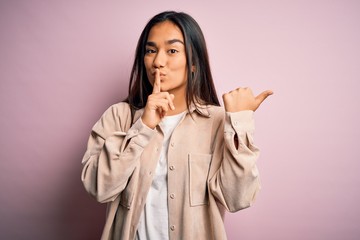 Young beautiful asian woman wearing casual shirt standing over pink background asking to be quiet with finger on lips pointing with hand to the side. Silence and secret concept.