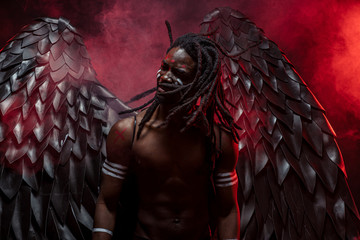 dark african angel with naked torso stand turning head with dreadlocks isolated over red smoky...