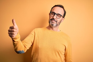 Middle age hoary man wearing casual sweater and glasses over isolated yellow background Looking proud, smiling doing thumbs up gesture to the side