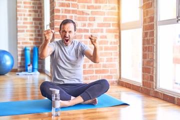 Middle age handsome sportman sitting on mat doing stretching yoga exercise at gym angry and mad raising fists frustrated and furious while shouting with anger. Rage and aggressive concept.