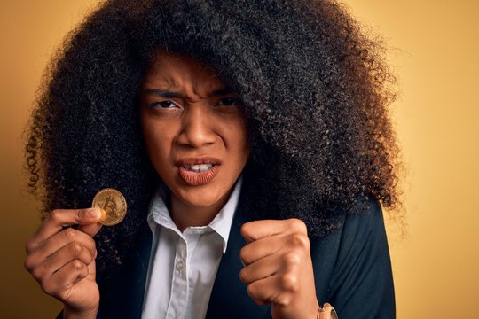 Young african american business woman with afro hair holding bitcoin over yellow background annoyed and frustrated shouting with anger, crazy and yelling with raised hand, anger concept