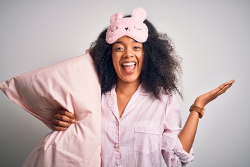 Young african american woman with afro hair wearing pajama and sleep mask holding pillow very happy...