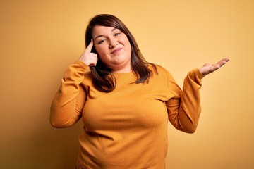 Beautiful brunette plus size woman wearing casual sweater over isolated yellow background confused and annoyed with open palm showing copy space and pointing finger to forehead. Think about it.