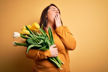 Beautiful plus size woman holding romantic bouquet of natural tulips flowers over yellow background bored yawning tired covering mouth with hand. Restless and sleepiness.