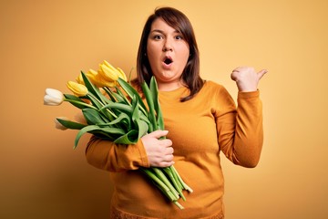 Beautiful plus size woman holding romantic bouquet of natural tulips flowers over yellow background Surprised pointing with hand finger to the side, open mouth amazed expression.