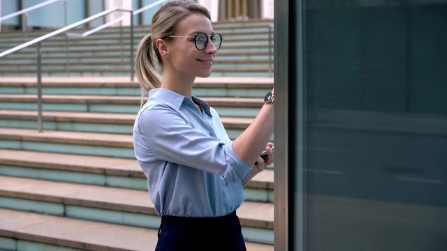 Millennial smiling female in classic glasses using big display for visitors standing at city street connected to public wireless on smartphone, happy woman tapping to screen of big interactive kiosk