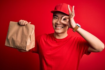 Young handsome chinese delivery man holding takeaway paper bag with food with happy face smiling doing ok sign with hand on eye looking through fingers