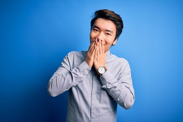 Young handsome chinese man wearing casual shirt standing over isolated blue background laughing and embarrassed giggle covering mouth with hands, gossip and scandal concept