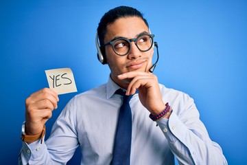 Young brazilian call center agent man holding reminder paper with yes message serious face thinking...
