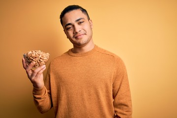 Young brazilian man holding bowl with healthy salty peanuts over isolated yellow background with a happy face standing and smiling with a confident smile showing teeth