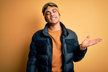 Young brazilian skier man wearing snow sportswear and ski goggles over yellow background smiling cheerful presenting and pointing with palm of hand looking at the camera.