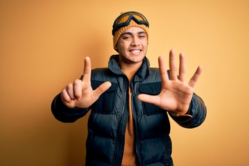 Young brazilian skier man wearing snow sportswear and ski goggles over yellow background showing and pointing up with fingers number seven while smiling confident and happy.
