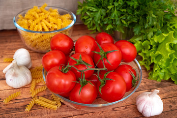 Bright, juicy, ripe tomatoes in a glass bowl on a wooden table , garlic in the foreground, greens and pasta in a glass jar in the background.