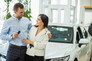 Fototapeta na wymiar portrait of married caucasian couple in car showroom. young man and woman stand posing against the backdrop of a new car, look at each other and hold keys from car in hands