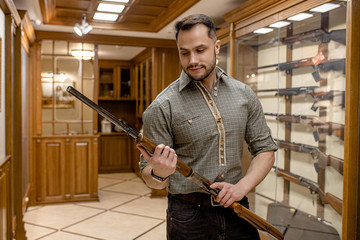 professional caucasian hunter looking for best rifle in guns store, he hold firearm in hands, want...