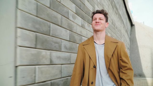 Stylish Beautiful Young Happy Caucasian Guy Goes along the City Wall and smiles. He is dressed in a Brown Long Coat. Close up - Powered by Adobe