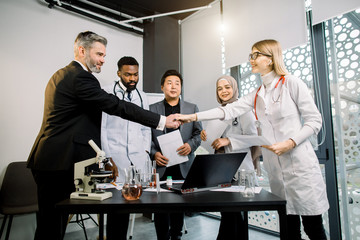 Team of doctors, scientists, pharmaceutical workers having meeting in modern lab with equipment. Caucasian man and woman shaking hands, after successful cooperation and results