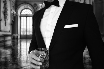 A close up view of a man in a black tuxedo holding a whiskey glass in a mansion. Great for use for a themed black tie event such as gatsbys mansion