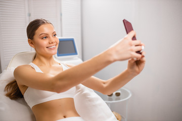 young female take selfie before hair removal procedure in beauty salon. young lady in white underwear smile at camera of smartphone