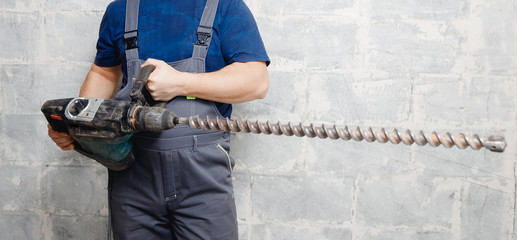 Male worker builder holds big drill background gray wall