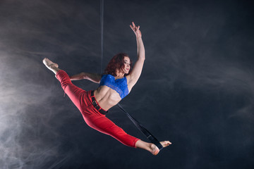 Female athletic, sexy and flexible aerial circus artist with redhead on aerial straps on black background