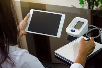 Mock up young woman look at blank screen of tablet computer with pressure guage and writing on notebook at home . Telemedicine, telehealth , e health concept .Over shoulder view.