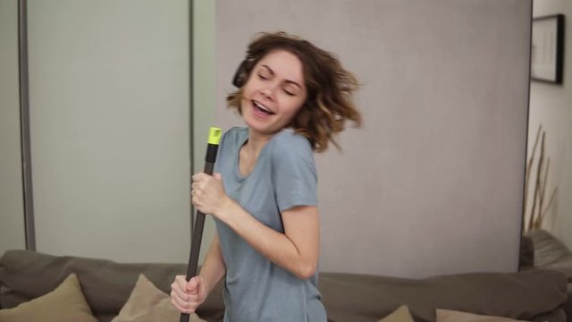 People, housework and housekeeping concept - happy woman in headphones with mop cleaning floor and dancing at home. Using swab pipe as a microphone. Grey couch on the background