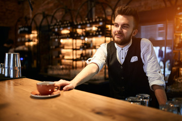 Portrait of smiling bearded stylish manstanding at table while having job in confectionary shop, offering a cup of tea. beverage Occupation concept. Copy space. close up side view photo