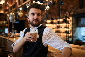Portrait of bearded young barman in stylish shirt standing, leaning on high bar table in cafeteria, free time, spare time, lifestyle