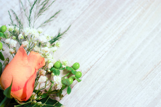 A peachy orange rose flower bud with white baby's breath (Gypsophila) and green hypericum berries against a white-washed wood background, with copy space
